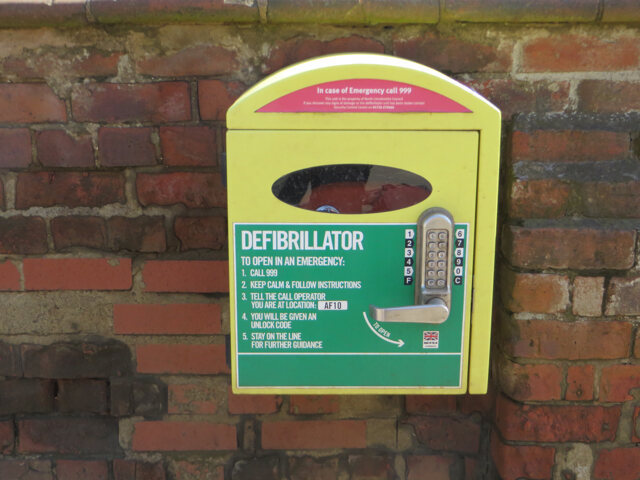 A yellow and green locked defibrillator cabinet mounted on a brick wall, with instructions to call 999